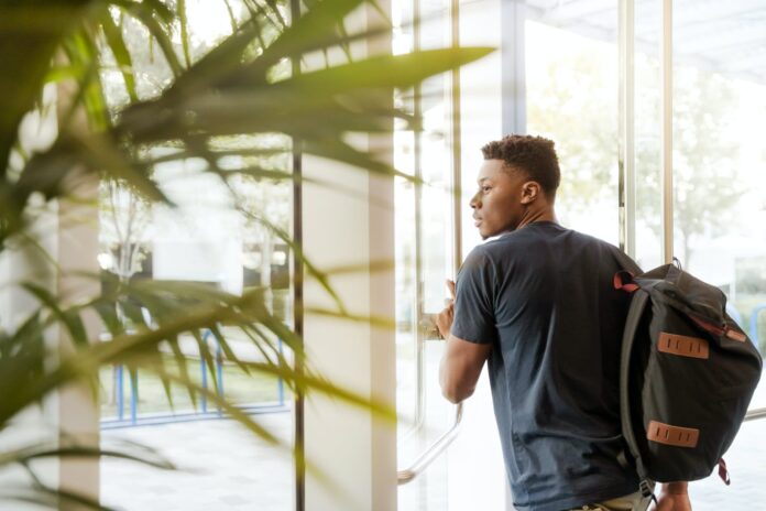 man looking outside window carrying black and brown backpack while holding his hand on window